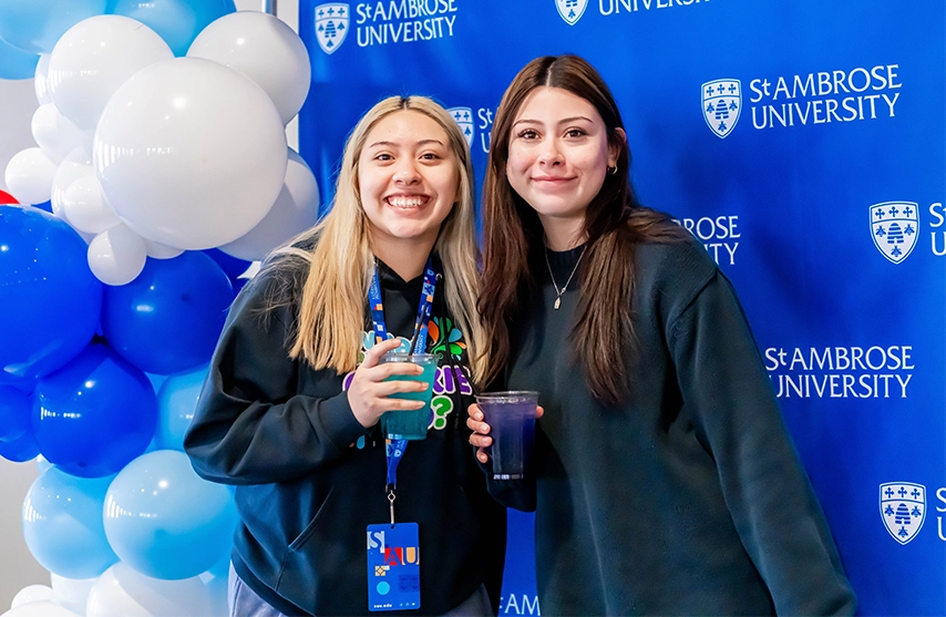 Two students holding an energy drink in front of a St. Ambrose sign
