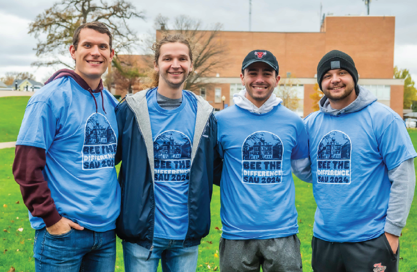Four smiling volunteers wearing blue “Bee the Difference SAU 2024” shirts stand together outdoors on a grassy area, posing for a group photo.