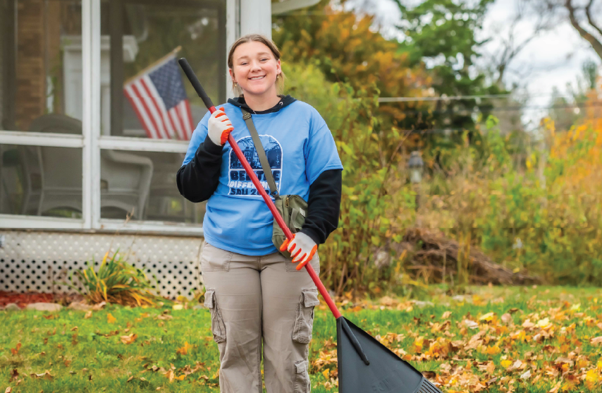 A smiling volunteer wearing a blue “Bee the Difference SAU 2024” shirt holds a rake while standing on a lawn covered in fall leaves.