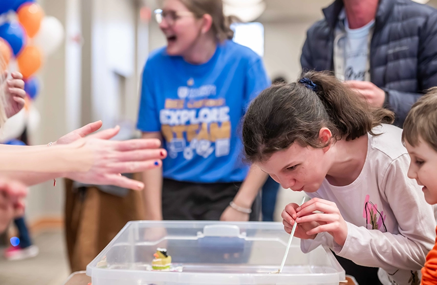 A young girl learns about science at St. Ambrose University's BEE Curious event as she blows through a straw to move objects in water. An adult woman with a Blue Bee Curious shirt smiles in the background as a crowd cheers the girl on.