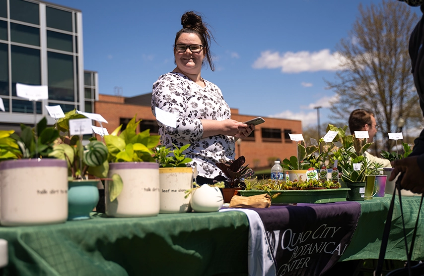Person standing behind a table of potted plants at an outdoor sustainability event smiling at camera.
