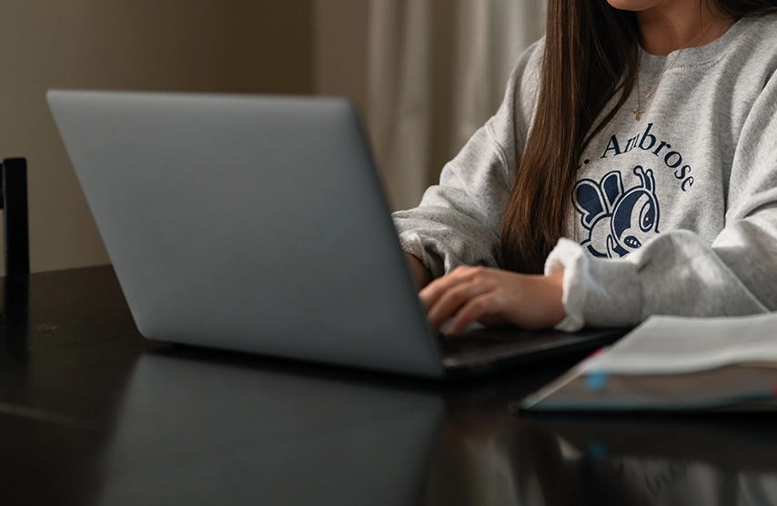A young woman works at a computer with an open book next to her.