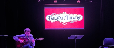 A student plays guitar on stage with a pink screen that reads 
