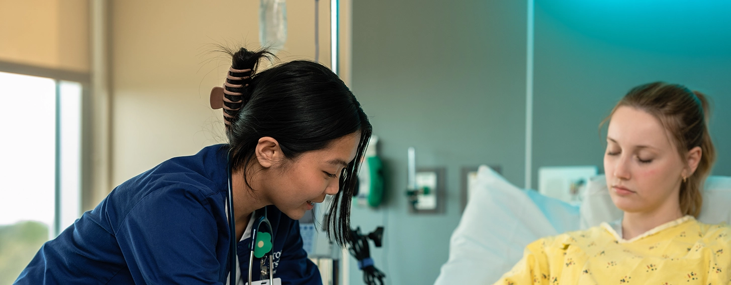 A student in scrubs leans over a patient in a yellow gown.