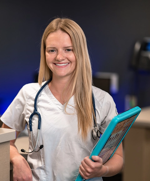A student in white scrubs with a blue clipboard smiles a the camera.