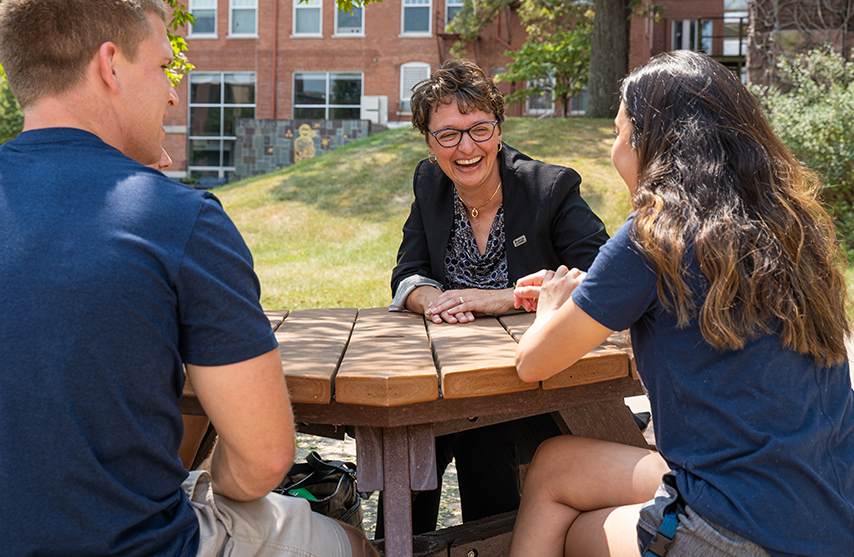 Amy Novak, EdD sits a table and laughs with two students dressed in dark blue.