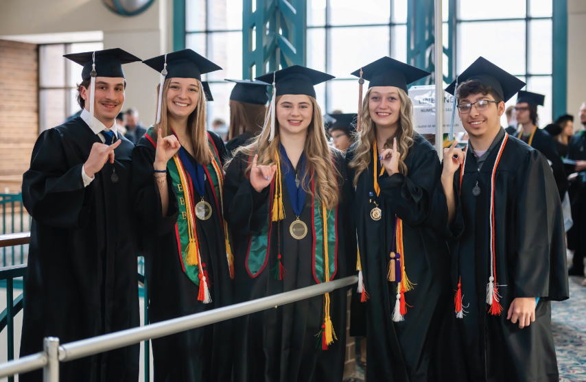Five students smiling together in caps and gowns with pinkies lifted (