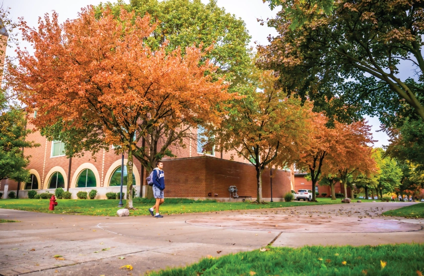 A student walking outside in front of orange and red trees.