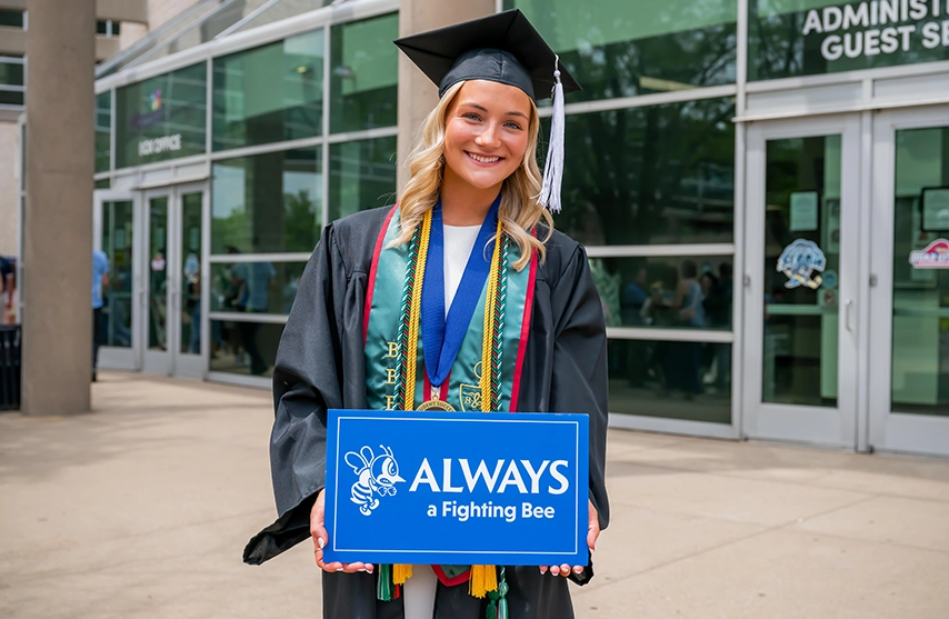 Ellyse Holubar stands in her graduation regalia outside of the 2026 SAU Spring Commencement. She holds a sign that reads, 