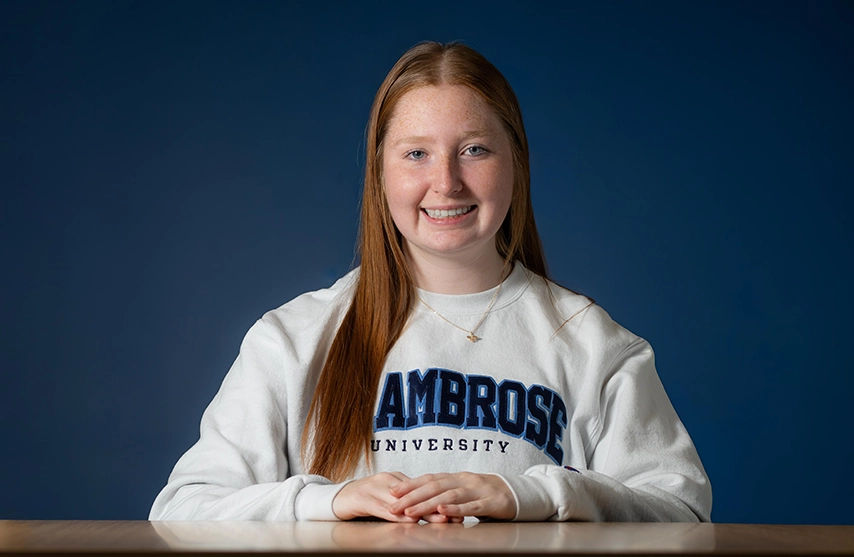 Jillian Aslip, '29, sits at a table and smiles into the camera.
