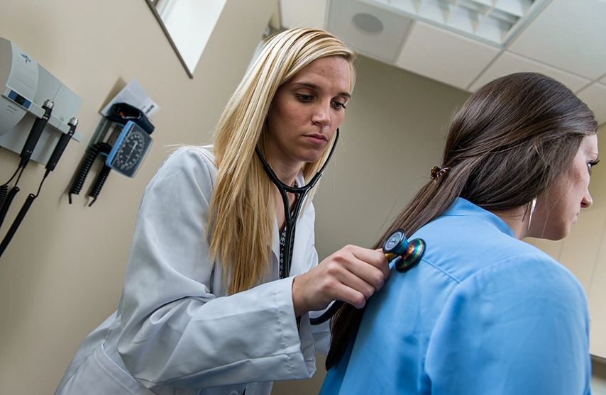 A student in a doctors' coat and stethoscope practices in their clinical.
