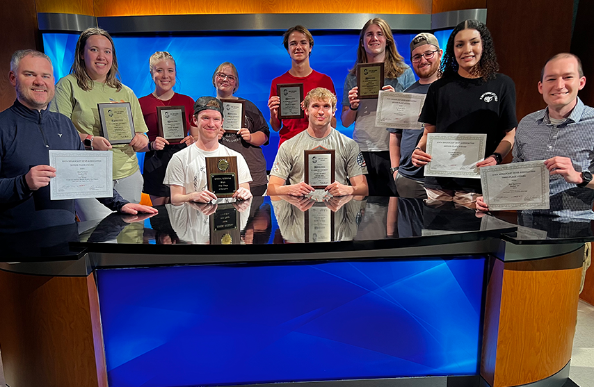 Students and faculty gather around a news desk and hold their many awards.