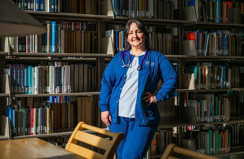 Kathy Franco stands in the St. Ambrose University Library in front of a shelf of books and smiles into the camera as a beam of light illuminates her.