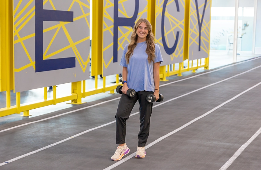 A student stands on an indoor track holding a dumbbell in each hand.