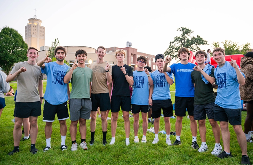 A group of St. Ambrose University students stands together on a campus lawn during an outdoor event, holding up their pinkies to show school spirit as 