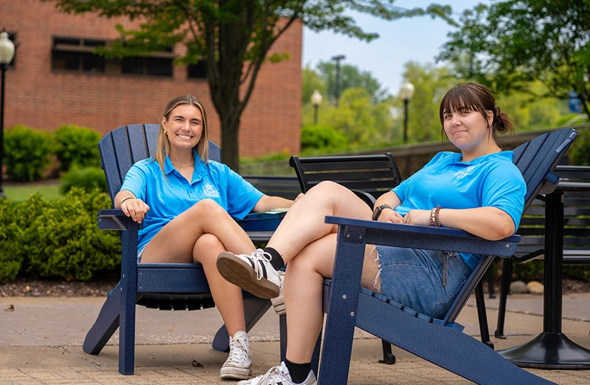 Students smile at the camera as they relax in two SAU blue adirondack chairs.