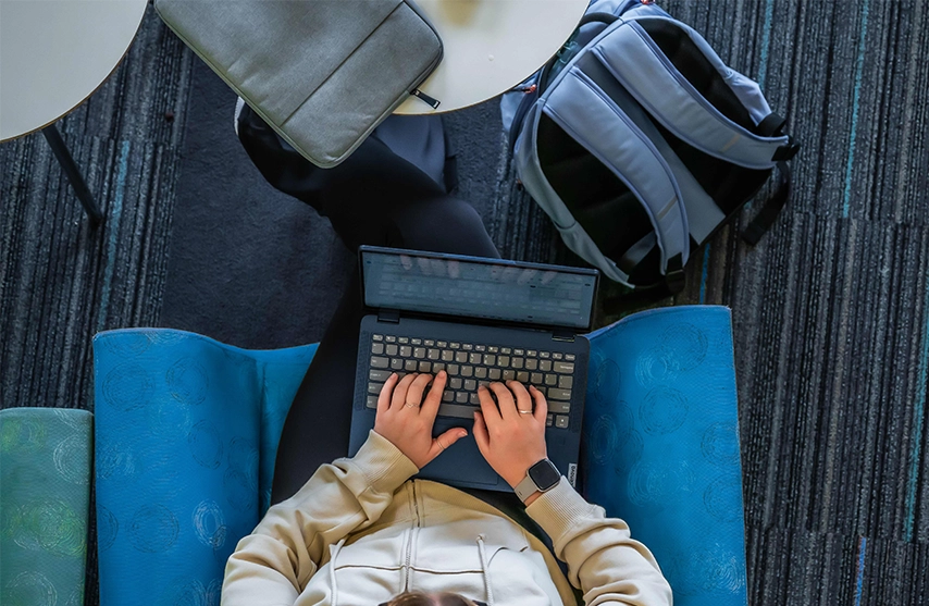 A student sits on a blue couch while typing on her laptop.