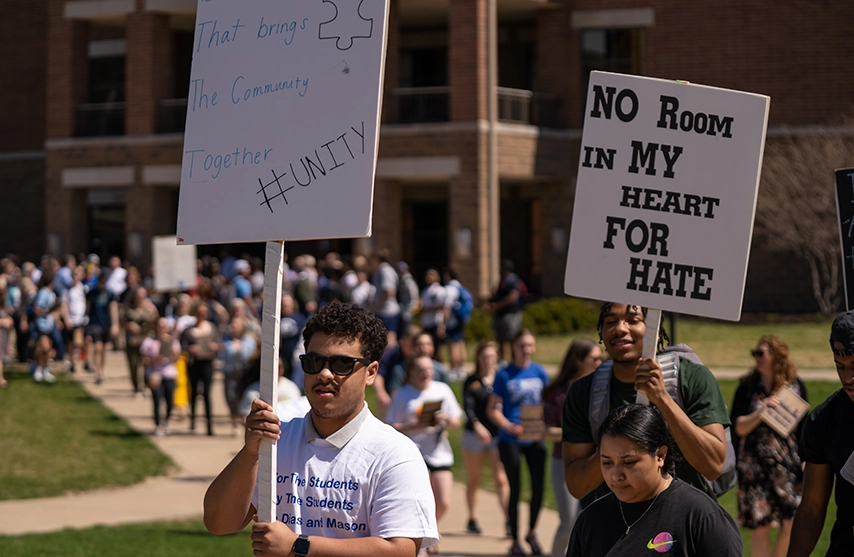 A long line of students stretches from the foreground to the background. A student in white holds up a sign that reads 