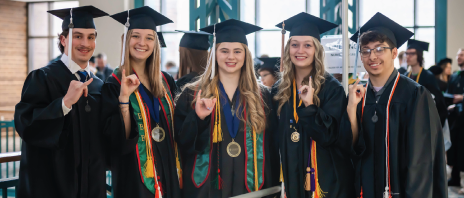 Five students smiling together in caps and gowns with pinkies lifted (