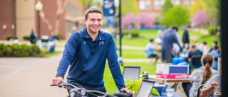 A student smiles at the camera during a club fair where he leans against a bike.