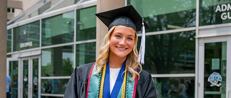 Ellyse Holubar stands in her graduation regalia outside of the 2026 SAU Spring Commencement. She holds a sign that reads, 