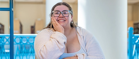 Nazaria Almanza-Gustafson, '26, smiles into the camera while sitting at a desk in the St. Ambrose University library.