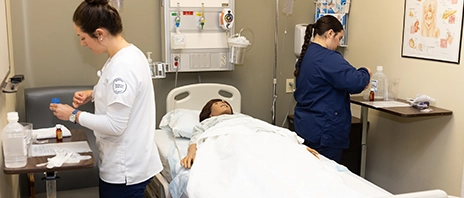 Two nursing students work together in a hospital room over a practice mannequin.
