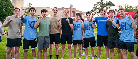 A group of St. Ambrose University students stands together on a campus lawn during an outdoor event, holding up their pinkies to show school spirit as 