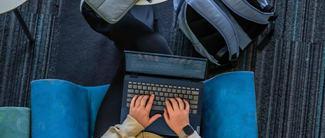 A student sits on a blue couch while typing on her laptop.