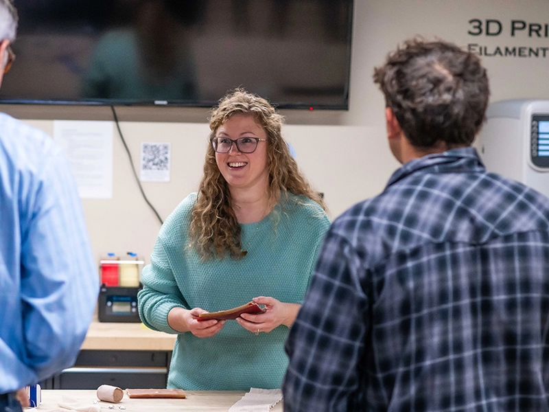 A young woman holds materials in her hands as she smiles at two people with their back to the camera.