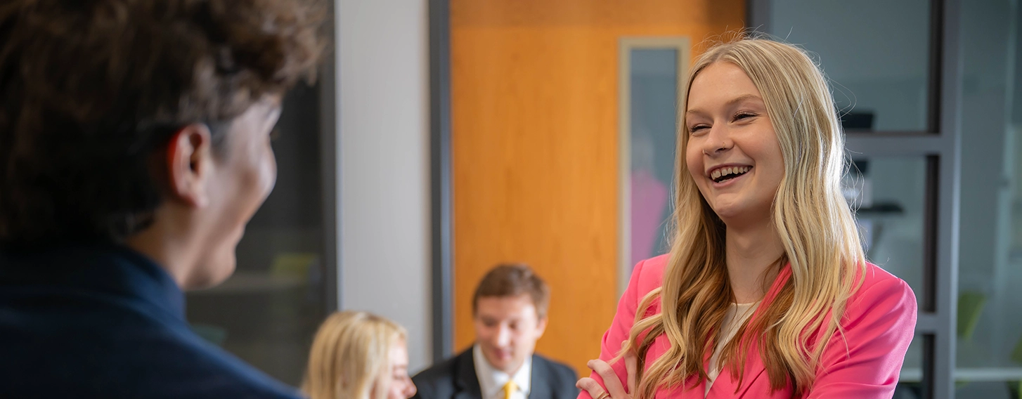 A BBA student stands in a pink suit with her arms crossed while laughing. Two other suited figures site in the background and work at a computer.